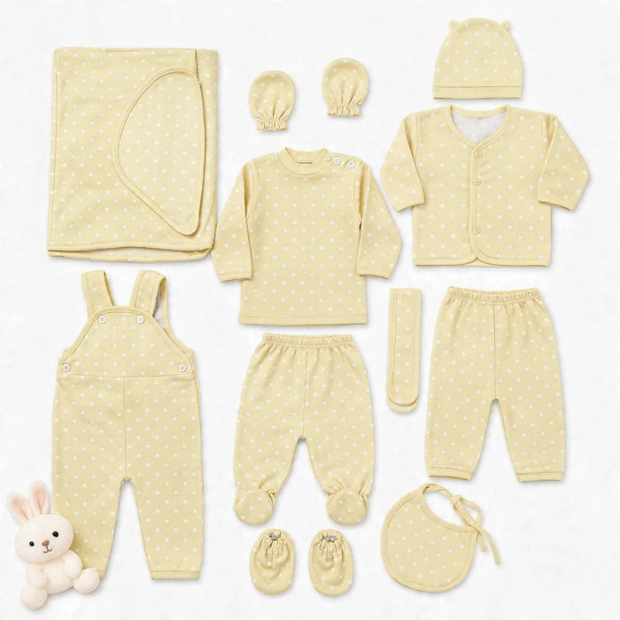 A coordinated baby clothing set laid out on a white background, including a long-sleeve top, footed pants, overalls, cardigan, hat, booties, bib, mittens, and a folded blanket. The set is a soft pale yellow with small white polka dots, and a small plush b
