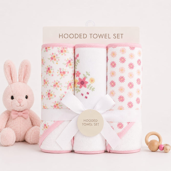 A three-pack of hooded baby towels rolled up and tied with a white ribbon, displayed on a clean white background with a plush pink bunny toy and a wooden teething ring prop.