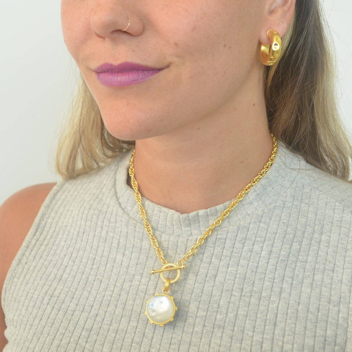 Woman wearing a gold necklace with a pearl pendant and matching earrings against a plain background