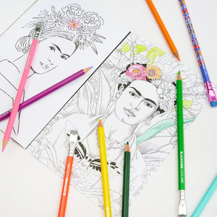 A styled flat-lay of coloring pencils and art supplies on a white surface, including a partially colored portrait drawing of Frida Kahlo with floral details, an uncolored black-and-white version, and several colored pencils in pink, purple, orange, yellow, green, blue, and teal.