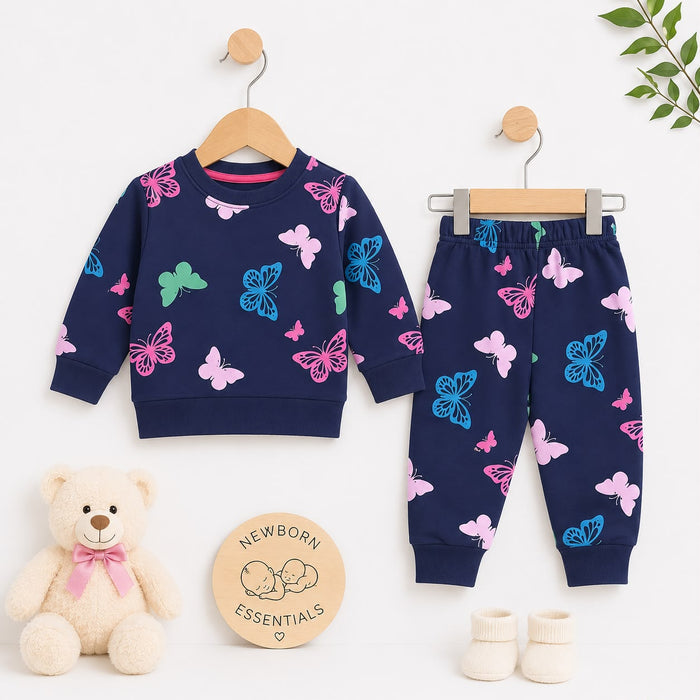 A baby clothing set displayed on wooden hangers against a white background, including a navy long-sleeve top and matching jogger pants with pink and blue butterfly prints. A cream teddy bear, a round wooden sign reading 'Newborn Essentials,' cream booties