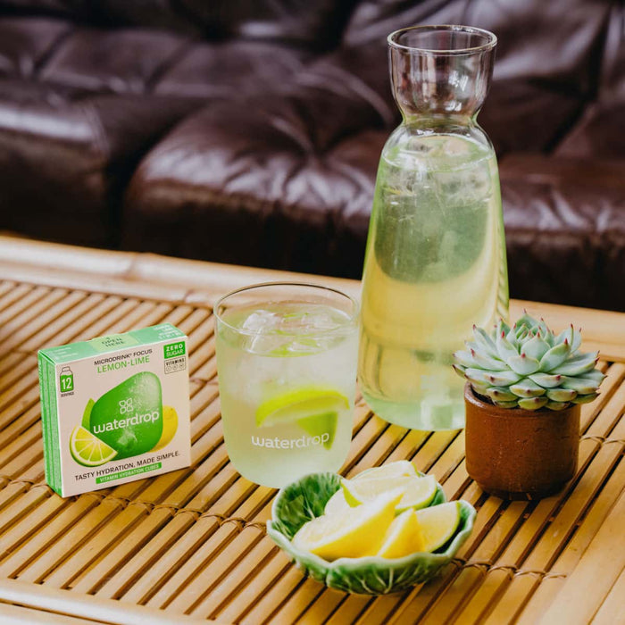 Glass of lemon lime drink with a carafe, lemon slices, & a small plant on a wooden table.