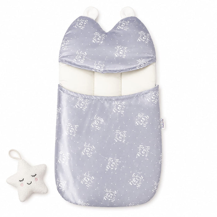 A baby footmuff in a light lavender-gray satin-like fabric with a white floral and star pattern, featuring a white inner lining and two white pom-poms. A small white plush star toy with embroidered facial features is placed to the left on a clean white ba