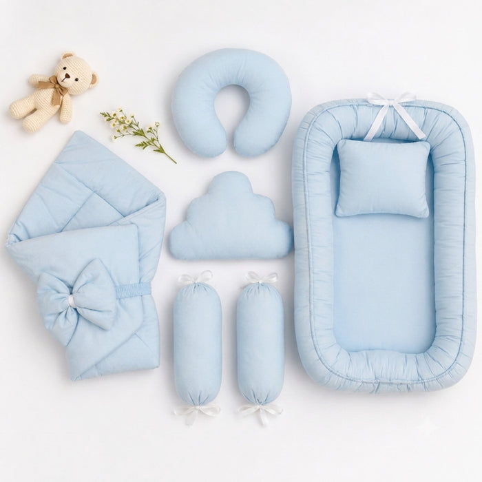 A coordinated baby bedding set arranged on a white background, including a bassinet with matching pillow, a folded blanket with bow, a U-shaped pillow, a cloud-shaped pillow, two cylindrical inserts, a small teddy bear prop, and a sprig of flowers.