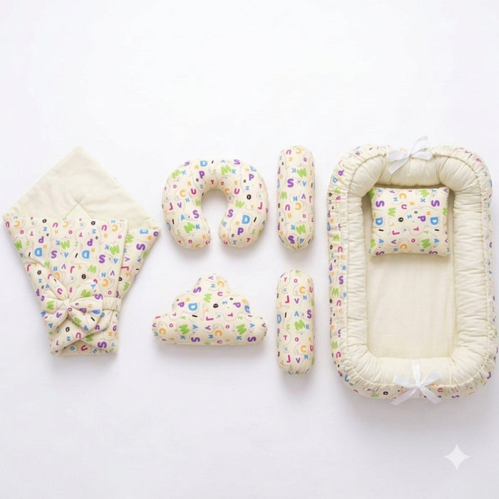 A baby bedding set laid out on a white background, including a quilted blanket with a colorful alphabet print and bow, a U-shaped pillow, two cylindrical pillows, a cloud-shaped pillow, and a bassinet cover with matching printed fabric and white ribbon ti