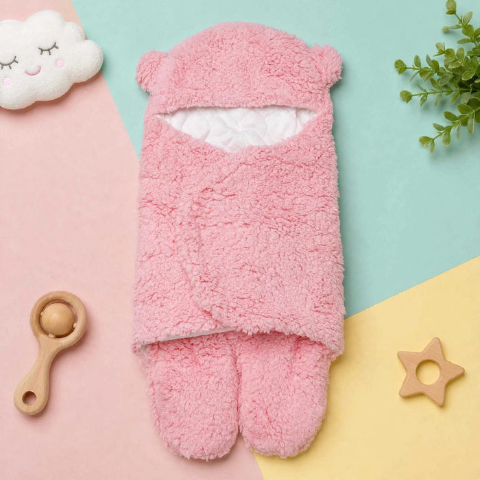 A pink plush baby foot warmer or bootie set laid flat on a pastel geometric background with a cloud toy, wooden rattle, greenery, and a star teether around it.