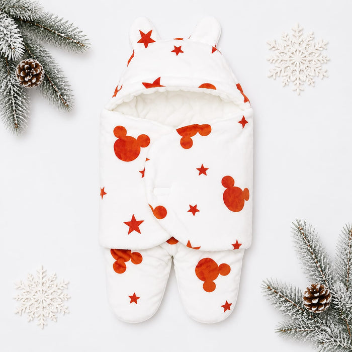 A baby footed swaddle blanket with a hood and ear details, featuring a white background with red Mickey Mouse head silhouettes and red stars. The swaddle is styled in a flat-lay with winter-themed props including pine branches, pinecones, and white snowfl