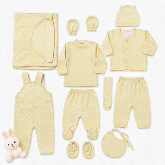 A coordinated baby clothing set laid out on a white background, including a long-sleeve top, footed pants, overalls, cardigan, hat, booties, bib, mittens, and a folded blanket. The set is a soft pale yellow with small white polka dots, and a small plush b