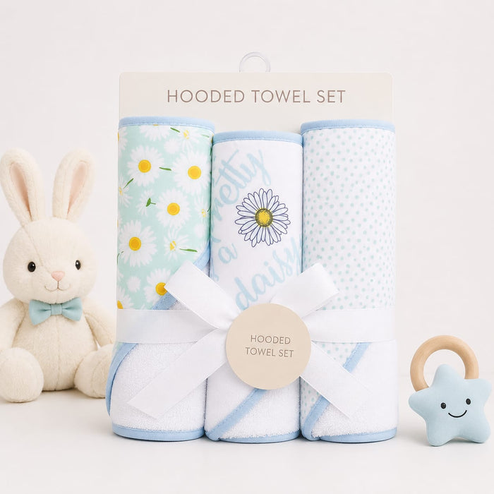 A hooded towel set displayed on a white background with a plush bunny toy on the left and a star-shaped teething ring on the right. The set includes three rolled towels: one with yellow daisy flowers on a mint green background, one with a daisy and the wo