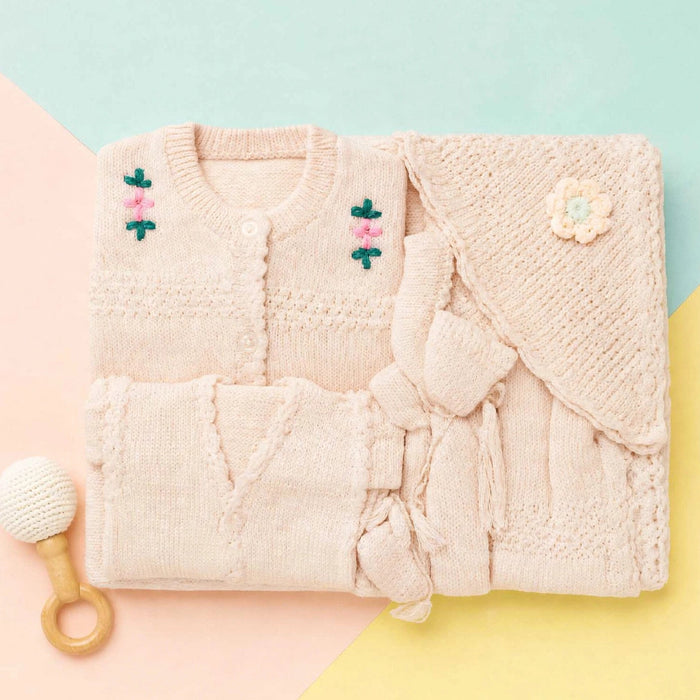 A folded knit baby outfit set in a soft blush pink, including a cardigan with embroidered flowers, a matching wrap or blanket with a flower applique, and a small wooden-handled baby rattle on a pastel geometric background.