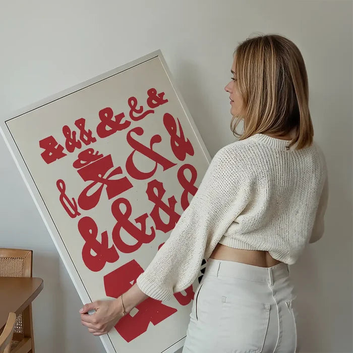 Woman holding a framed "Ampersands Colour Typography Print" with red ampersands on a cream background.