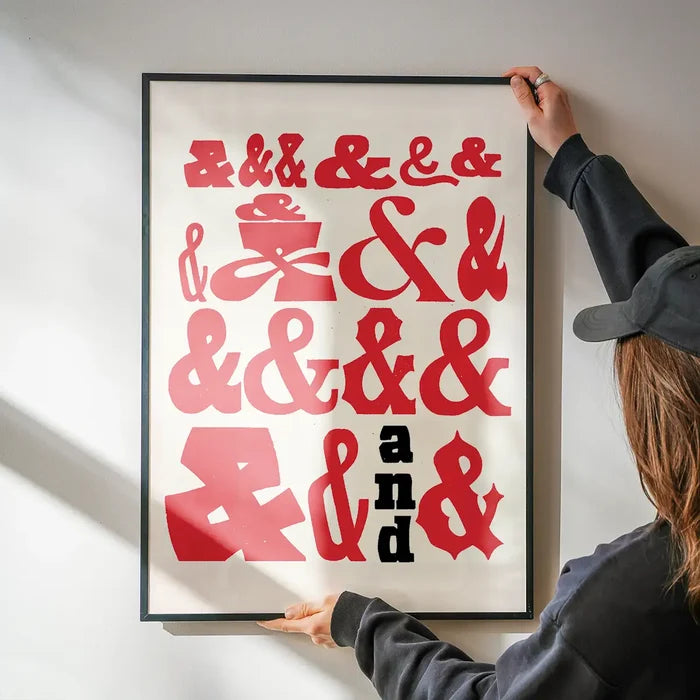 Red ampersands in various fonts on a white print, held by hands in a black frame.