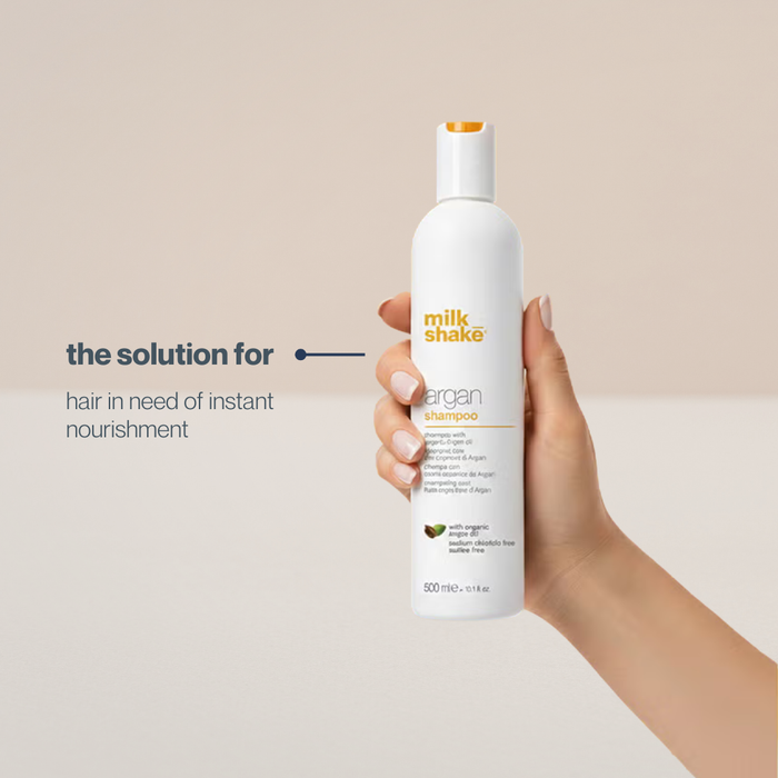Hand holding a bottle of milk_shake Argan Shampoo against a beige background