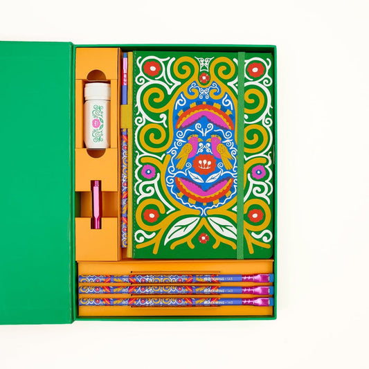 Colorful Frida Kahlo stationery gift set arranged inside a green box on a white background. The set includes a decorative notebook with Frida Kahlo's artwork, several matching patterned pencils, a white sharpener with a floral and vine illustration, and a pink point guard.