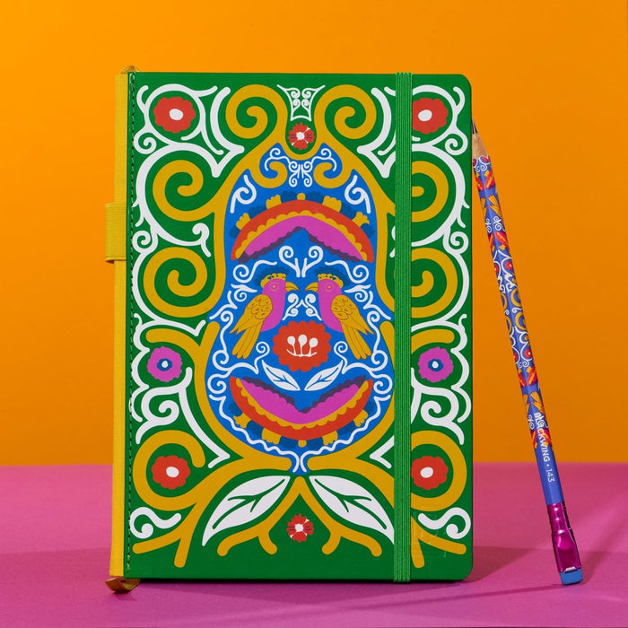 A hardcover notebook with a vibrant, multicolored Frida Kahlo design featuring birds and swirl patterns on a green background, shown next to a matching patterned pencil on a pink and orange background.