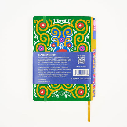 Back of a hardcover notebook featuring a vibrant green cover decorated with a colorful Frida Kahlo art pattern. The notebook is wrapped in a blue paper band with product details, barcode, and a QR code, and is shown with a colorful matching pencil on the right side holster.