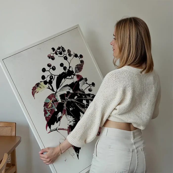 Woman holding a framed botanical print with black berries, colorful leaves, and graffiti art details.