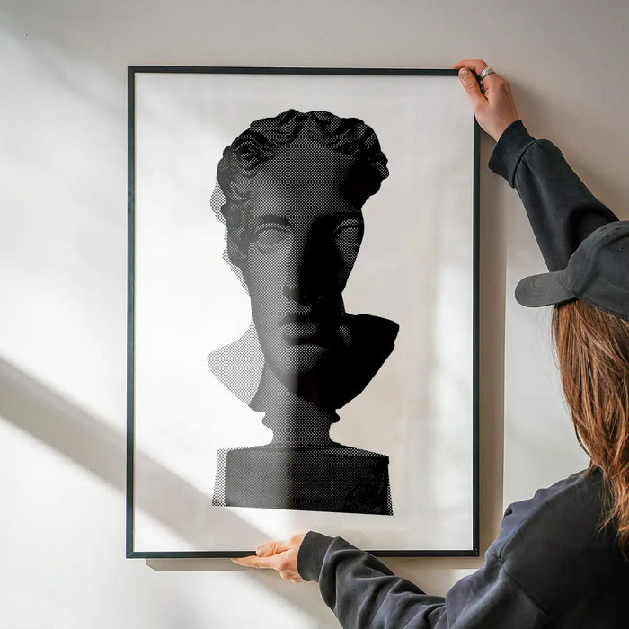 Framed black and white graphic print of a classic Roman bust, held by hands on a white wall.