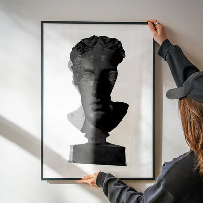 Framed black and white graphic print of a classic Roman bust, held by hands on a white wall.