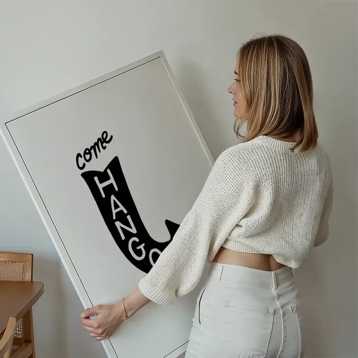 Woman holding a large white framed "Come Hang Out" typographic art print with black text.