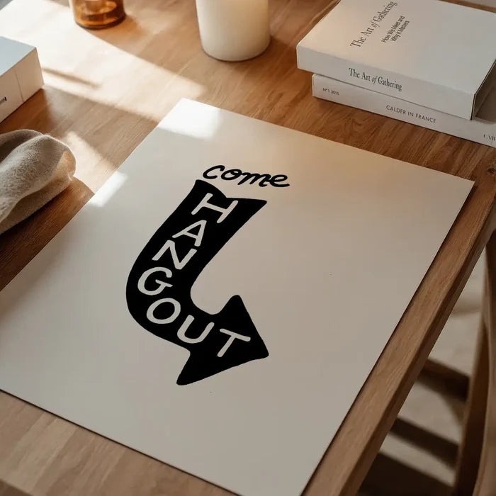 White art print with "Come Hang Out" in a black arrow shape, on a wooden table.