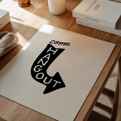 White art print with "Come Hang Out" in a black arrow shape, on a wooden table.