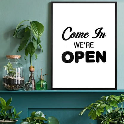 Black framed "Come In We're Open" typographic slogan print on white paper, displayed on a teal shelf with plants.