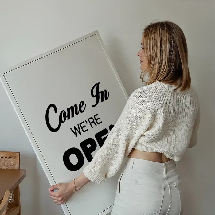 Woman holding a large white framed "Come In We're Open" slogan print with black typography.