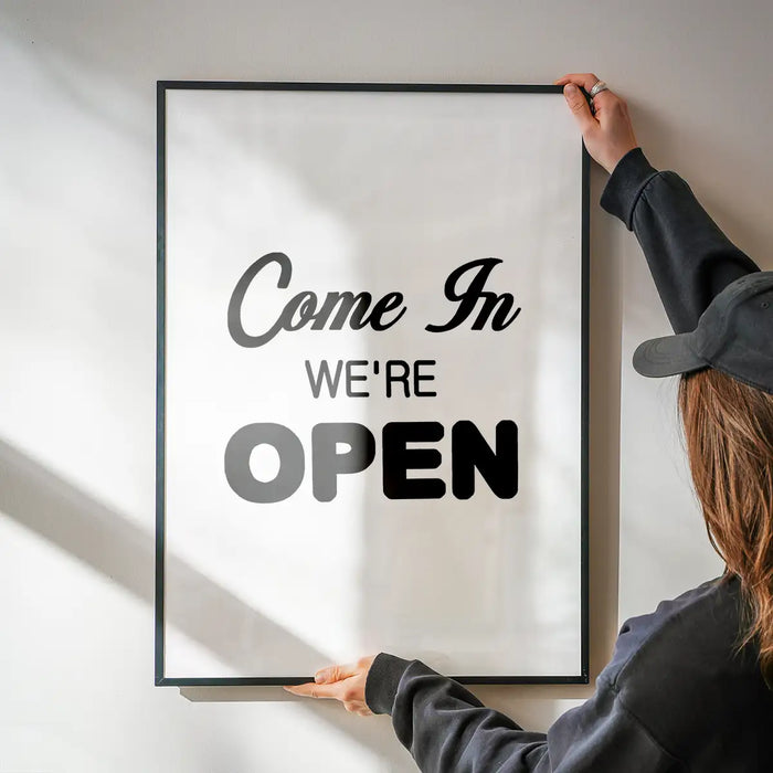 Black framed "Come In We're Open" slogan print on white archival art paper, held by a person.