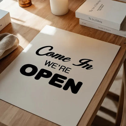 White typographic art print with "Come In WE'RE OPEN" in black text on a wooden table.