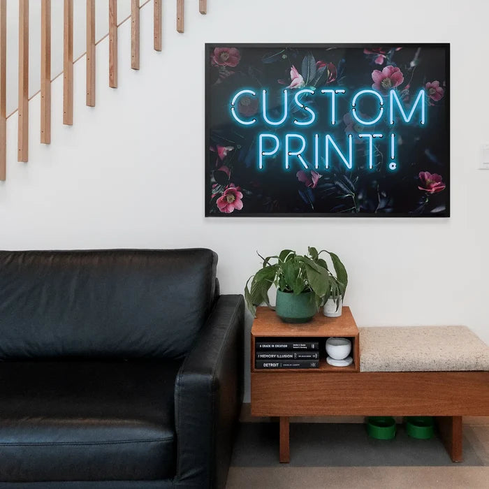 Blue neon sign print with "CUSTOM PRINT!" text and pink floral background, framed above a black sofa.
