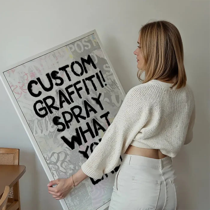 Woman holding a framed custom graffiti stencil print with "CUSTOM GRAFFITI! SPRAY WHAT YOU" in black spray paint.