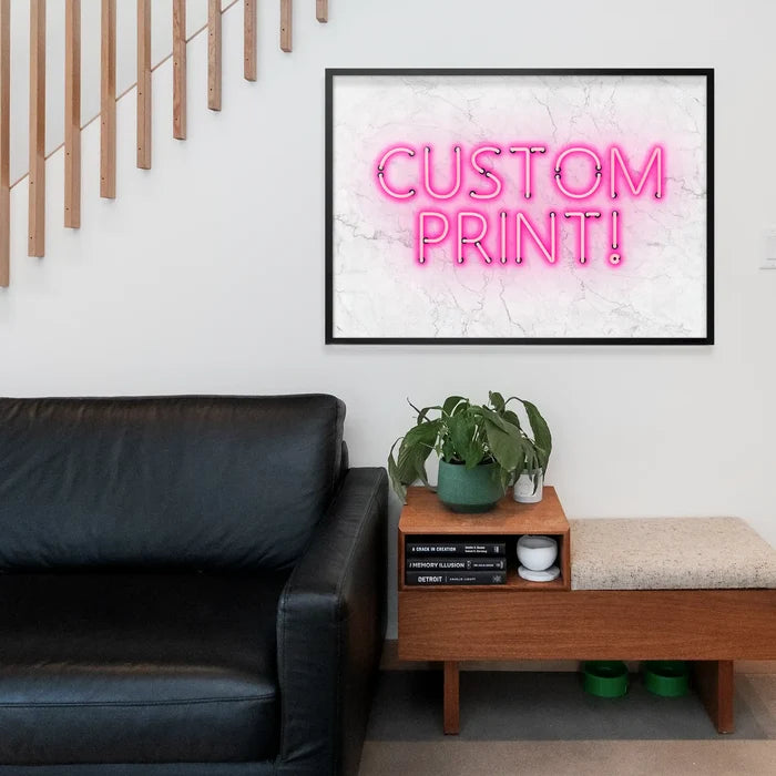 Pink "CUSTOM PRINT!" text on a white marble background, framed in black, hanging above a sofa.