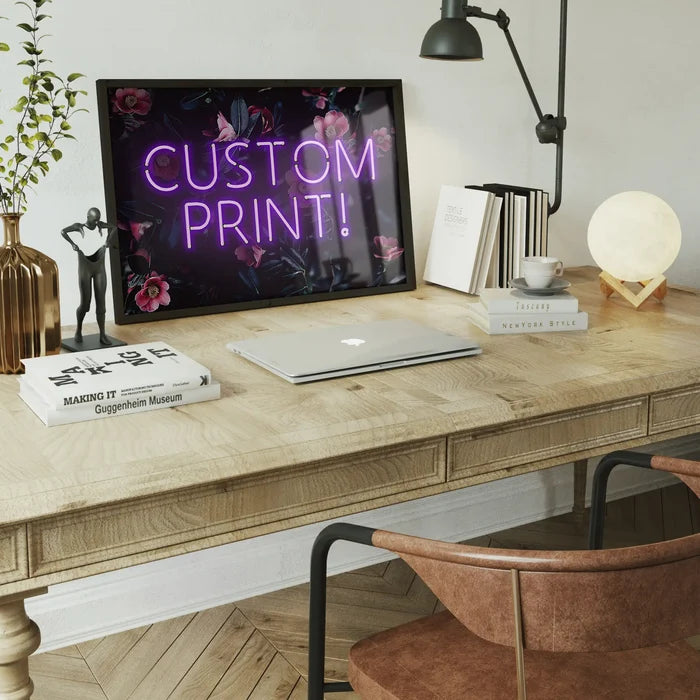 Purple neon "CUSTOM PRINT!" sign on a dark floral background, framed on a wooden desk with a laptop.