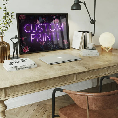 Purple neon "CUSTOM PRINT!" sign on a dark floral background, framed on a wooden desk with a laptop.