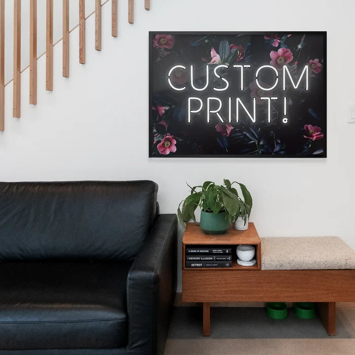 White neon "CUSTOM PRINT!" text on a dark floral background print, framed above a black leather sofa.