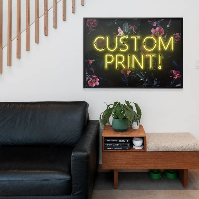 Yellow neon "CUSTOM PRINT!" text on a dark floral background print, framed above a black sofa.
