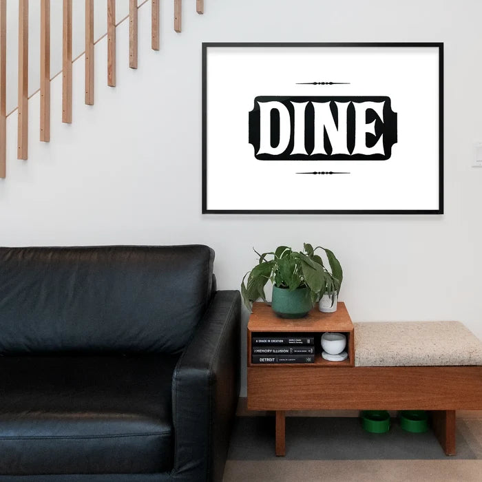 Black and white "DINE" typographic print in a black frame, hanging above a wooden bench and black sofa.