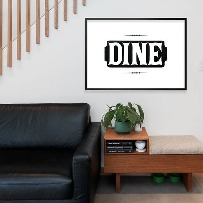 Black and white "DINE" typographic print in a black frame, hanging above a wooden bench and black sofa.