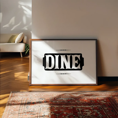 Framed "DINE" typographic print on white archival paper with ornate details, leaning on a wall above a patterned rug.