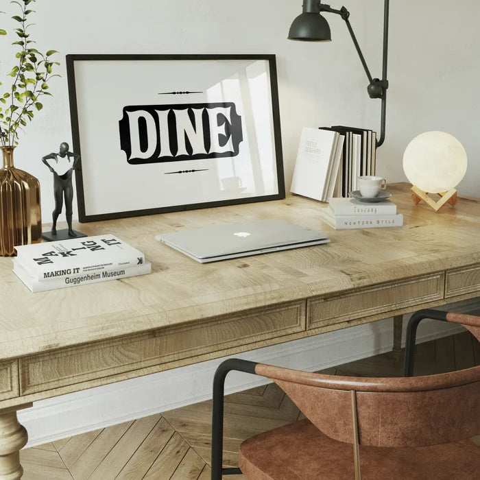 Black and white "DINE" typographic print in a black frame on a wooden desk with a laptop and decor.