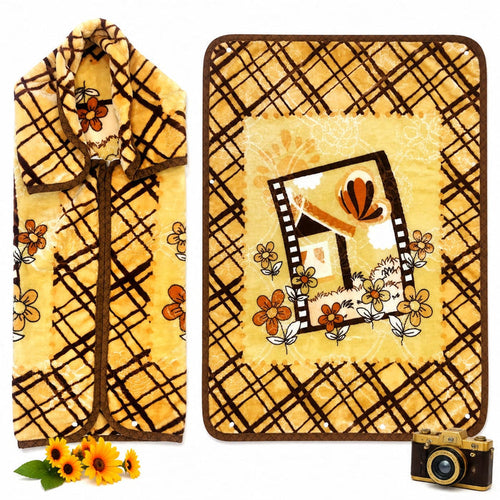 A yellow fleece blanket with a brown plaid pattern and a central floral house design, shown folded on the left and laid flat on the right. The blanket features a warm yellow base with brown grid lines, orange flowers, and a small house illustration in the