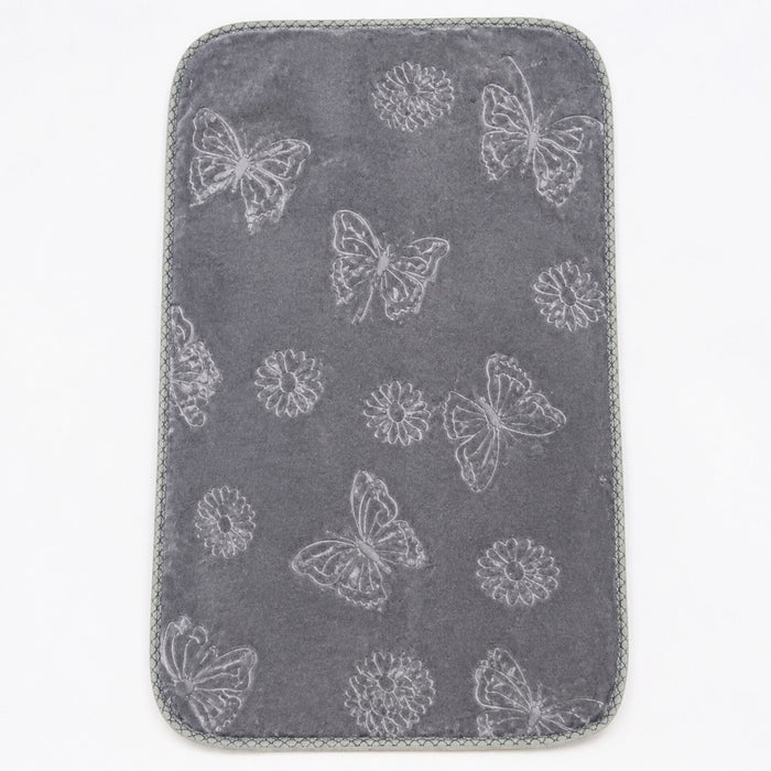 A dark gray rectangular mat with rounded corners, featuring a light gray embroidered pattern of butterflies, dandelions, and flowers, finished with a gray braided border, shown on a white background.