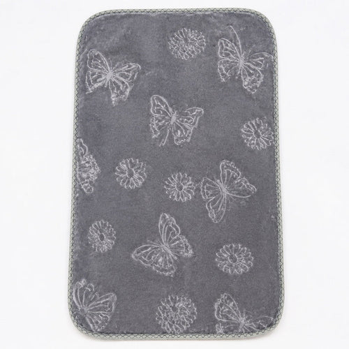 A dark gray rectangular mat with rounded corners, featuring a light gray embroidered pattern of butterflies, dandelions, and flowers, finished with a gray braided border, shown on a white background.