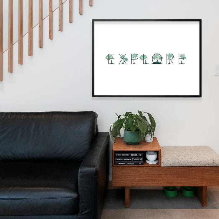 Black framed "EXPLORE" typographic print with green palm tree letters on white archival paper, above a black sofa.