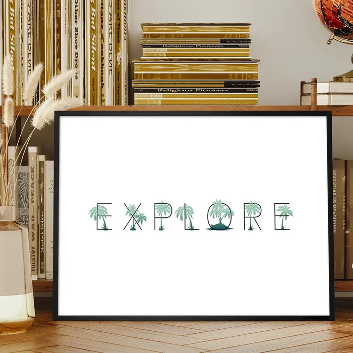 Black-framed "EXPLORE" print with green palm tree typography on white archival art paper, displayed on a wooden shelf.