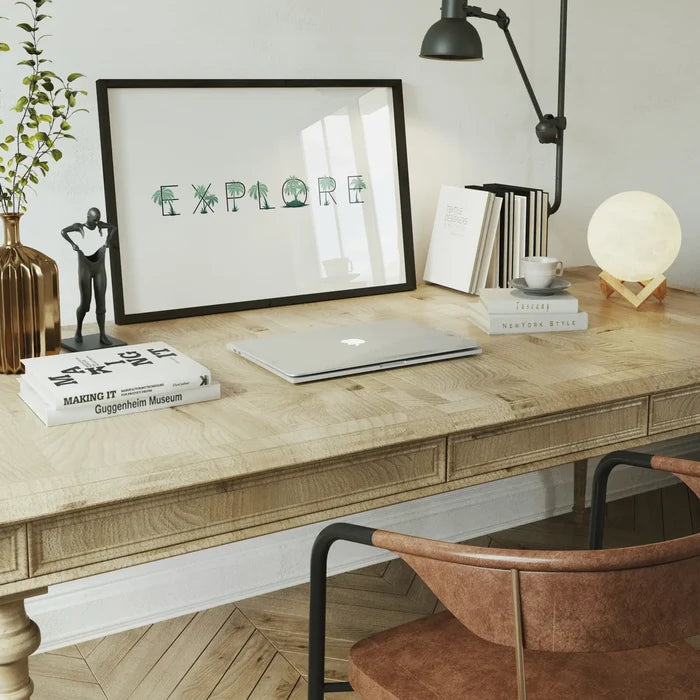 Framed "EXPLORE" typographic print with green palm tree letters on a wooden desk.