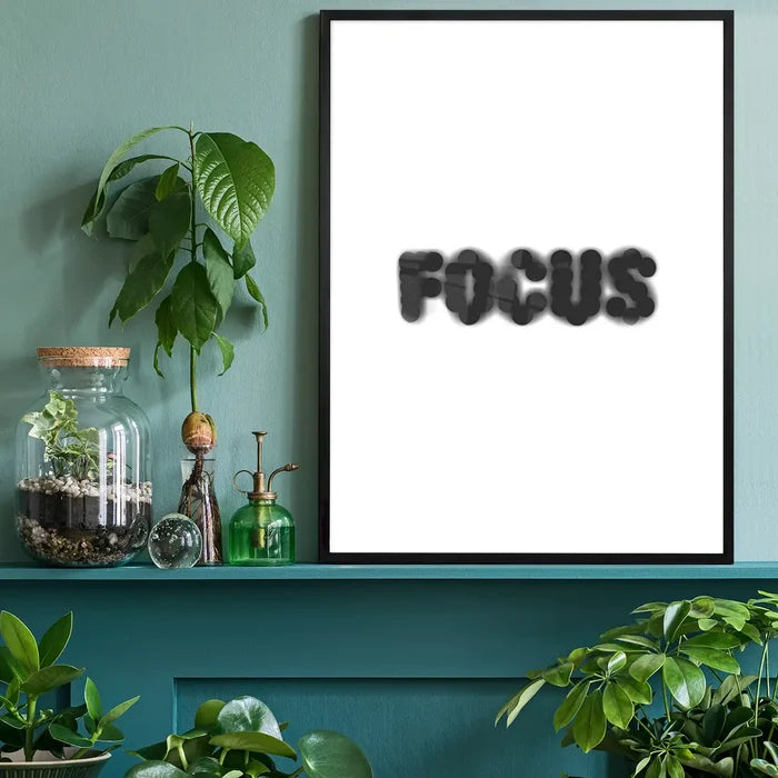 Black framed "FOCUS" typographic print on white archival paper, displayed on a green shelf with plants.