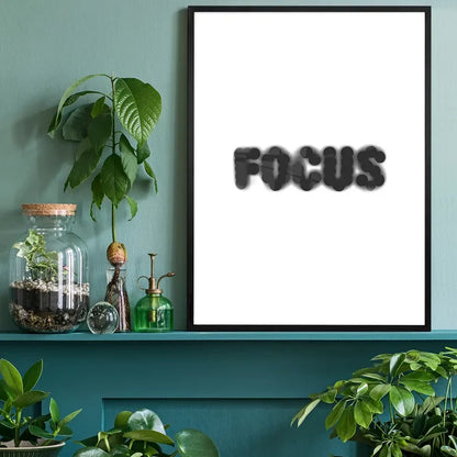 Black framed "FOCUS" typographic print on white archival paper, displayed on a green shelf with plants.