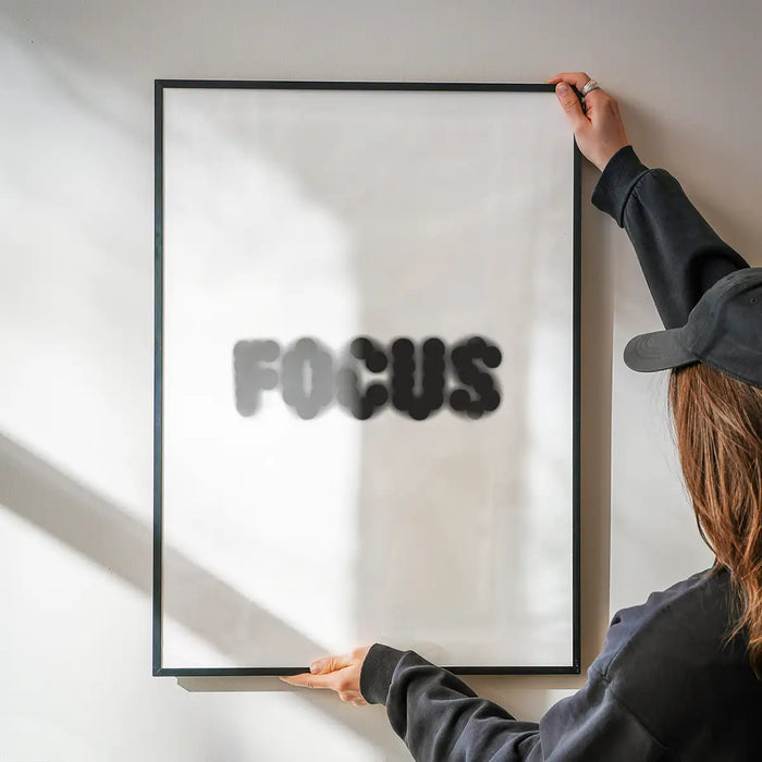 Person hanging a framed "FOCUS" quote print with blurred black text on white archival art paper.
